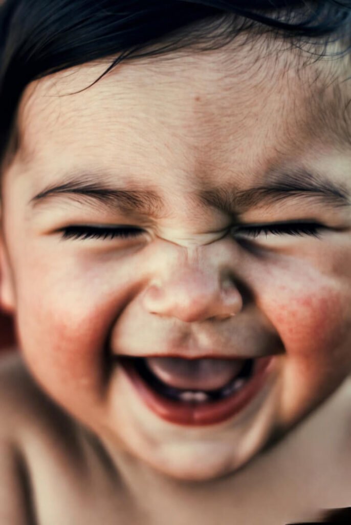 a baby mid-giggle, eyes squinted, cheeks puffed out. Soft lighting, slightly vintage tone.